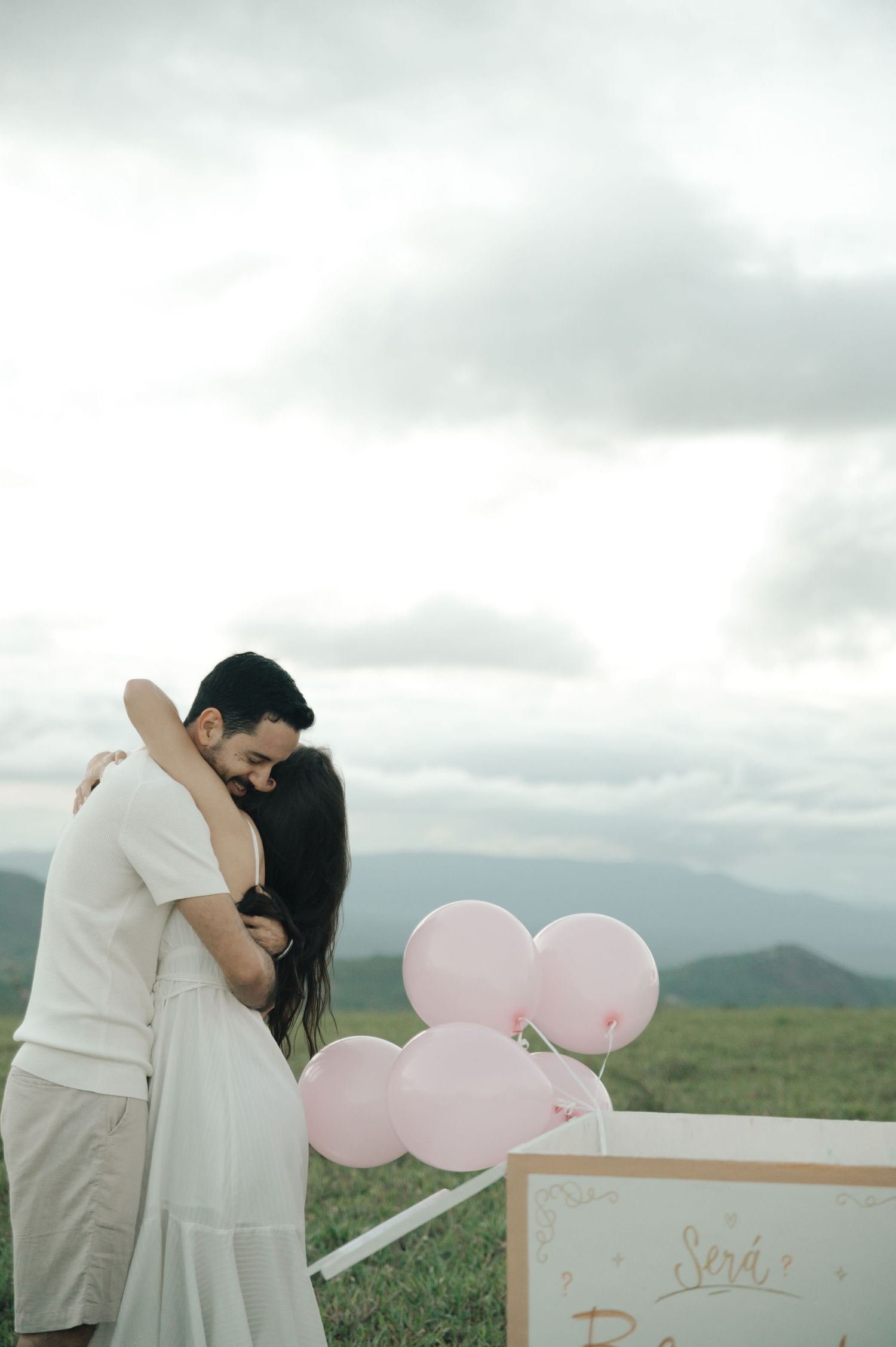 A couple embracing during a gender reveal with pink balloons and a sign on a scenic hilltop.