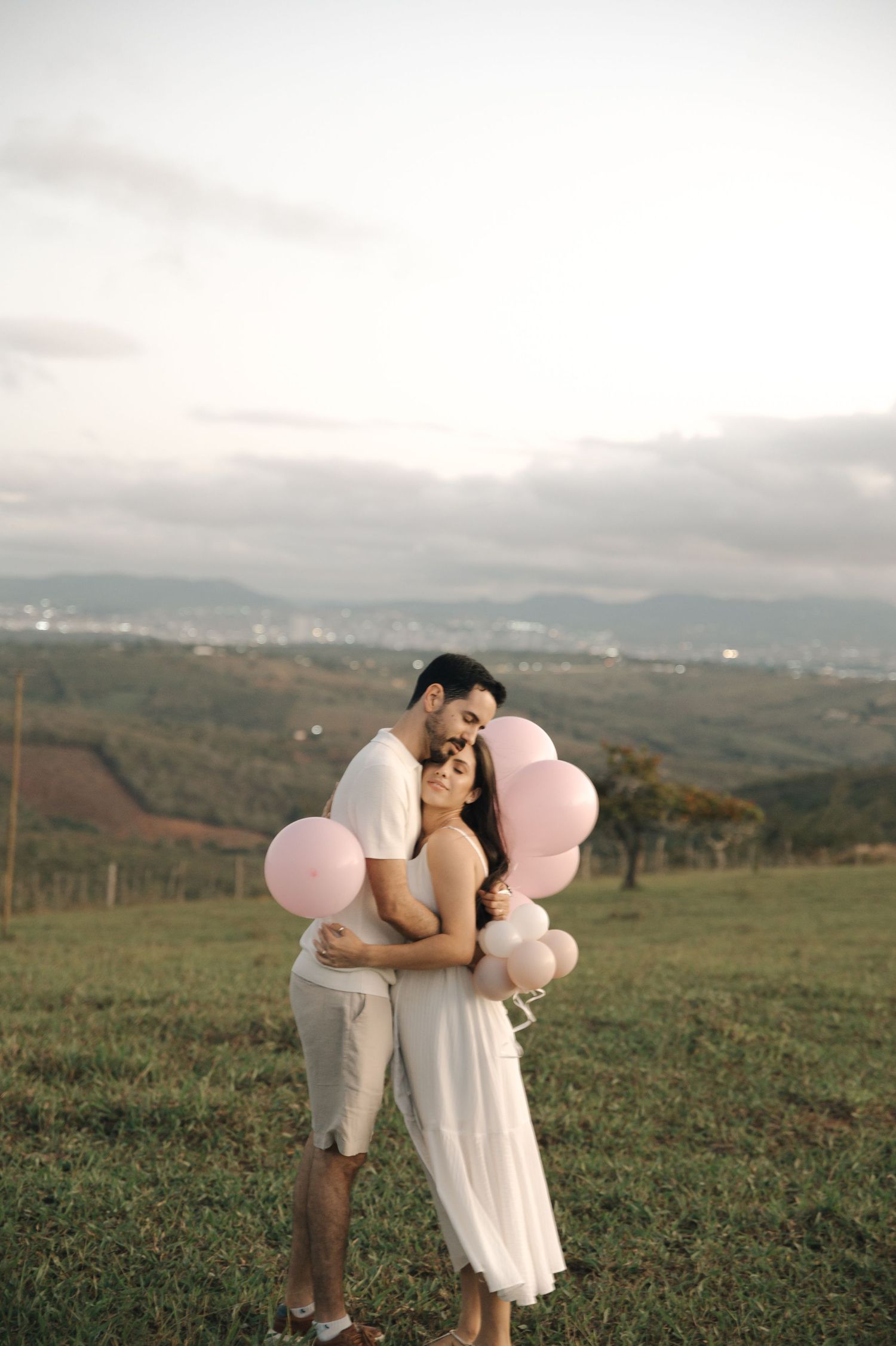 A couple embraces while holding pink balloons on a green hillside at sunset.