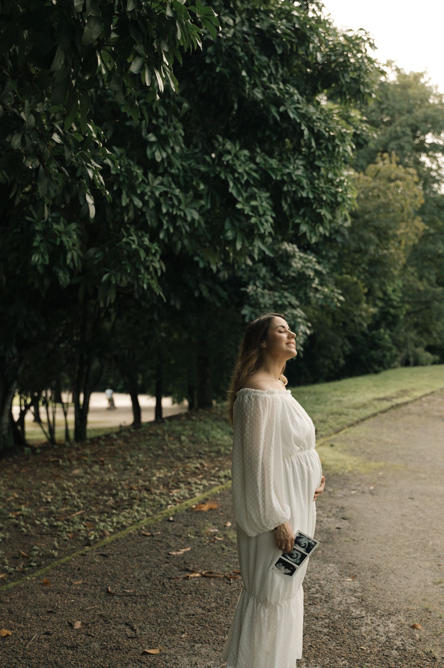 Pregnant woman in white dress holding ultrasound photos in a lush green park.