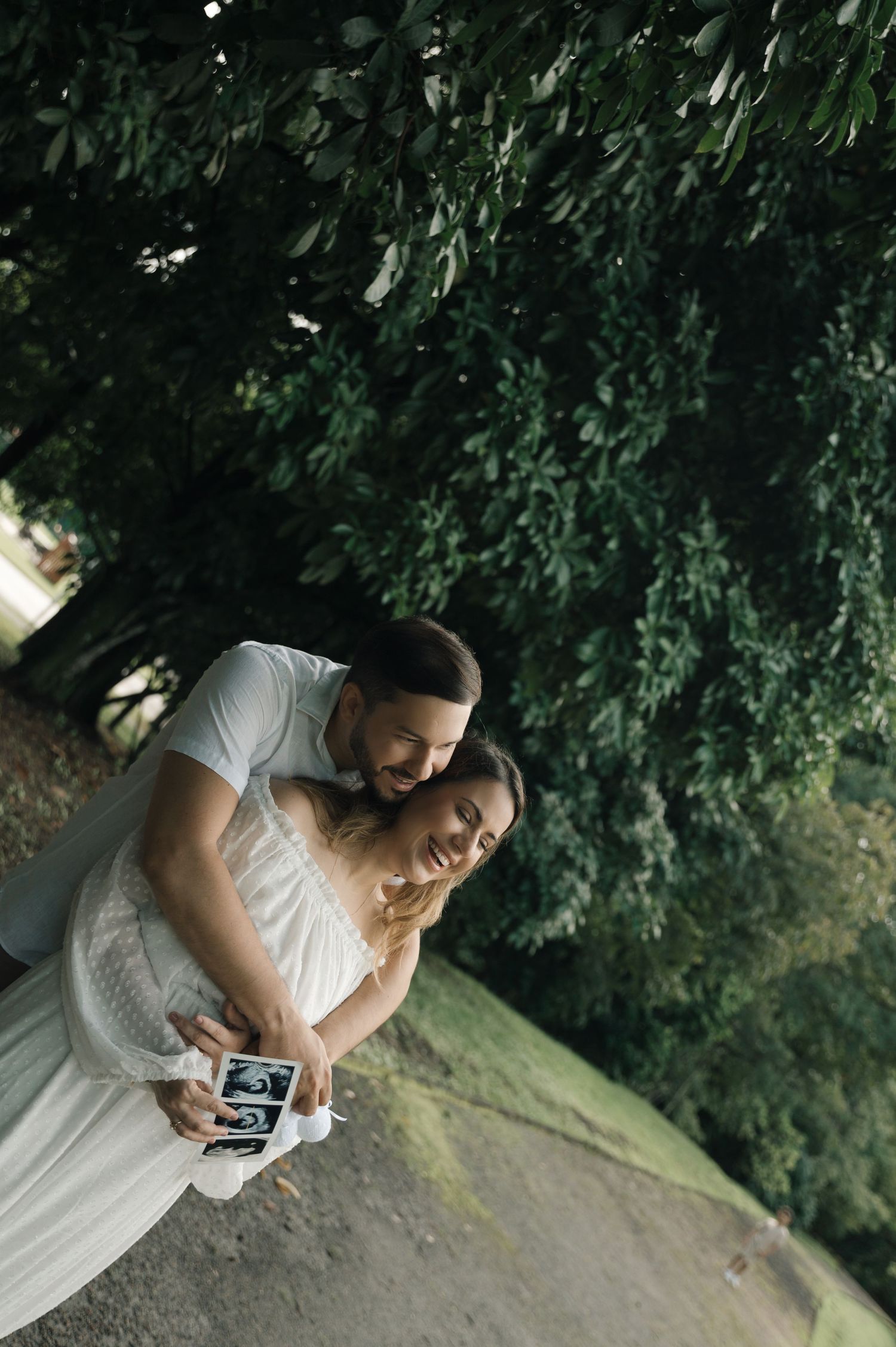 Couple embracing outdoors under lush green trees, holding ultrasound photos, celebrating pregnancy announcement.