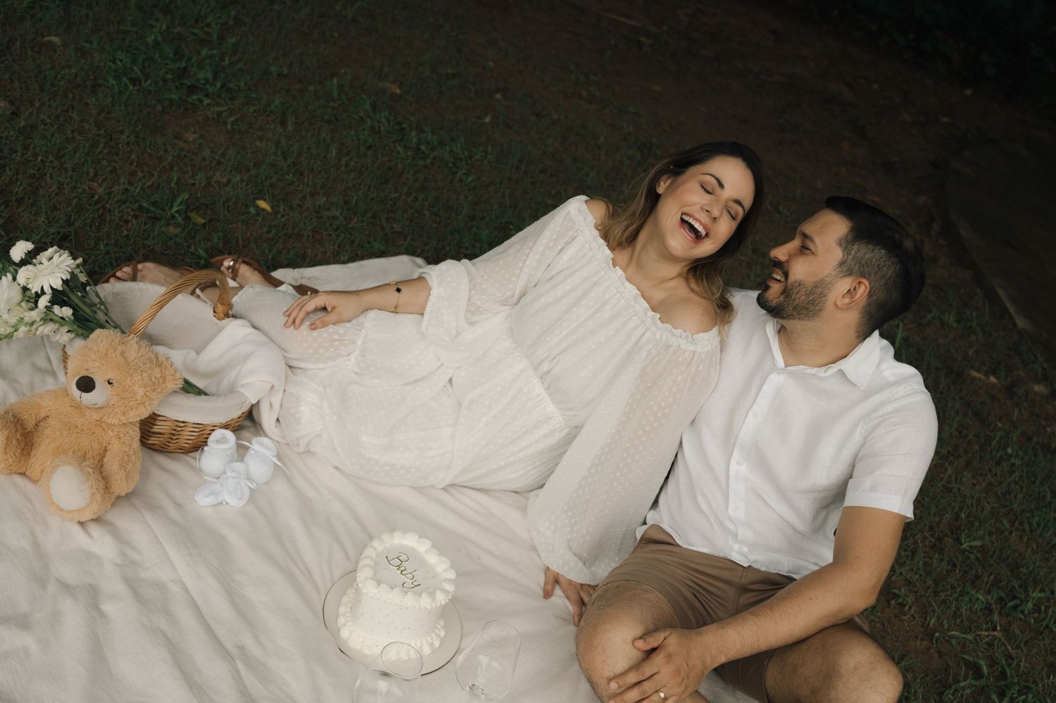 Couple laughing on white picnic blanket with teddy bear and cake outdoors.