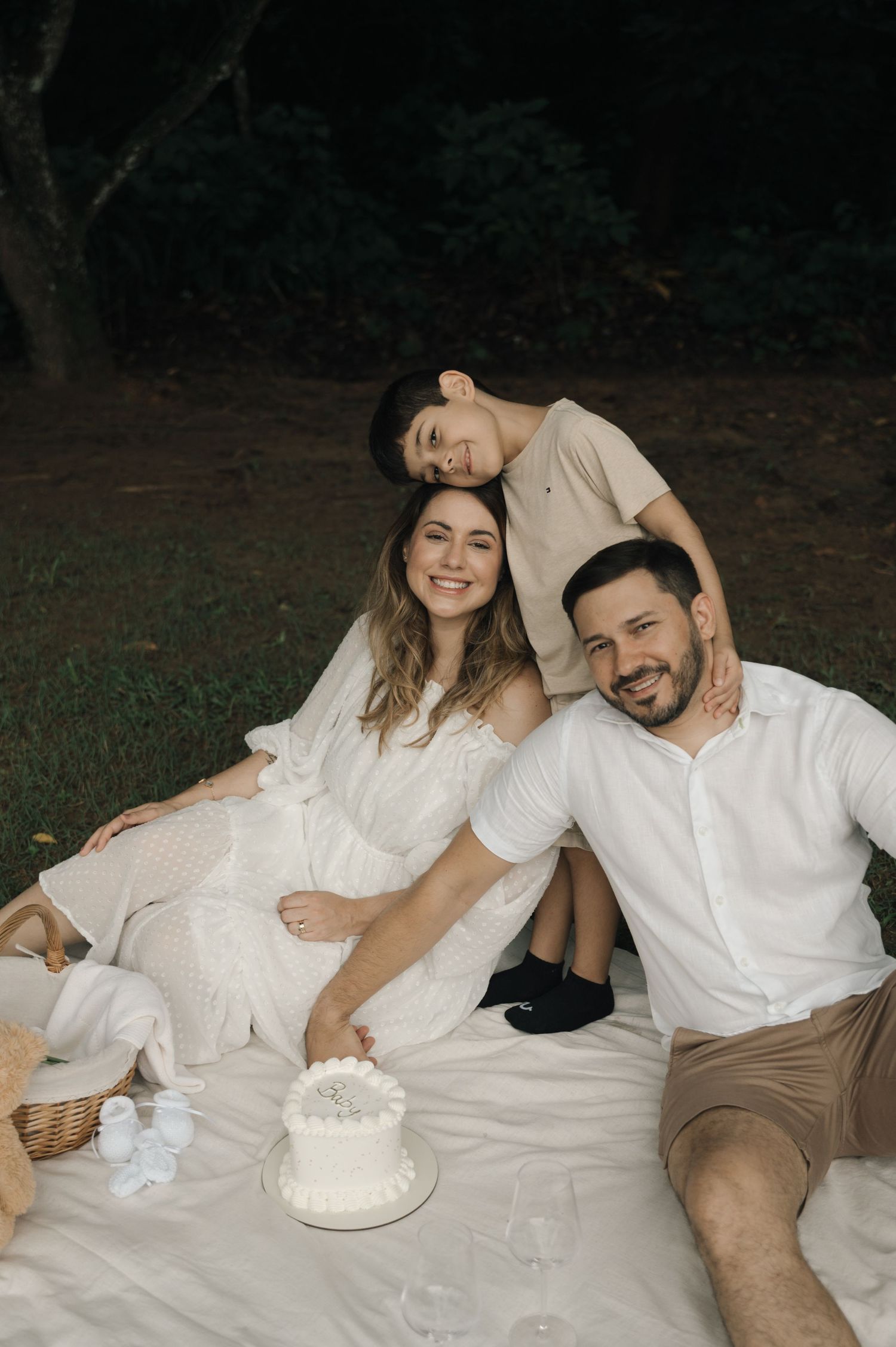 Family of three sitting on white blanket outdoors with white cake, smiling during a garden picnic celebration.