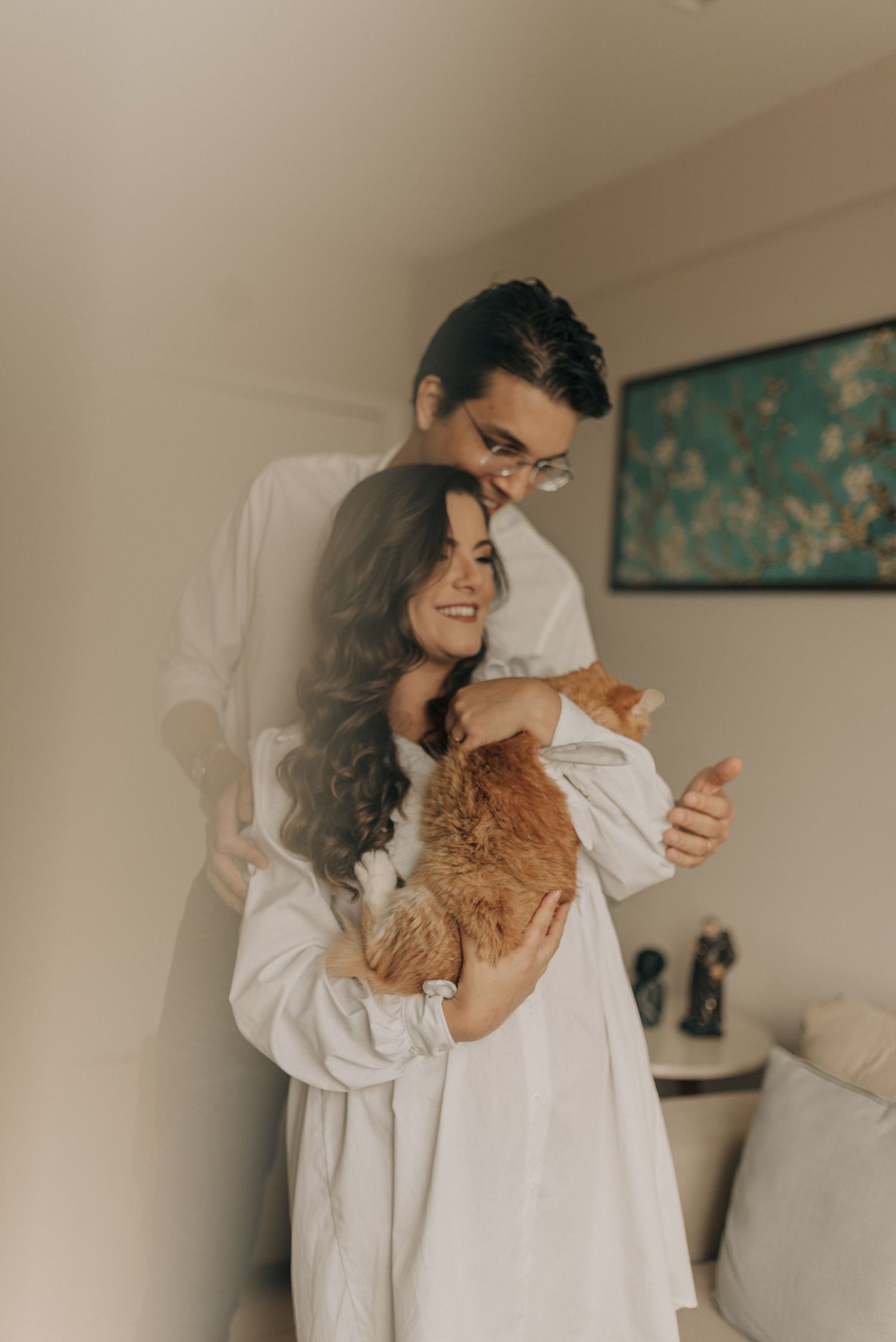 Couple in white clothing embracing with orange cat in cozy home setting.
