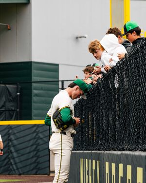 Oregon Ducks baseball player signing autographs for young fans leaning over the outfield fence after a game.