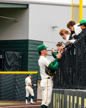 Baseball player in green uniform signs autographs for fans through a fence at a college baseball stadium.