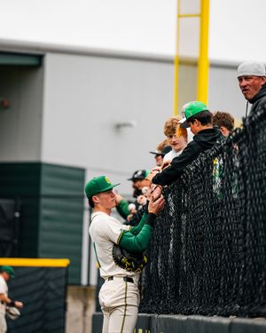 Baseball player in green uniform interacts with fans through a fence at an outdoor ballpark.