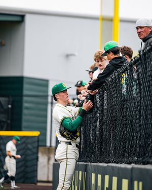 Baseball player in green uniform signs autographs for fans through a fence at a stadium.