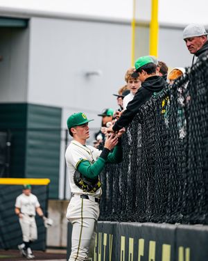 Oregon Ducks baseball player in green uniform interacts with fans through chain-link fence at PK Park stadium.