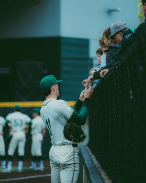Baseball player in green uniform signs autographs for young fans leaning over the outfield fence.