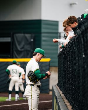 Baseball player in green and white uniform signs autographs for fans along the dugout fence during a game.