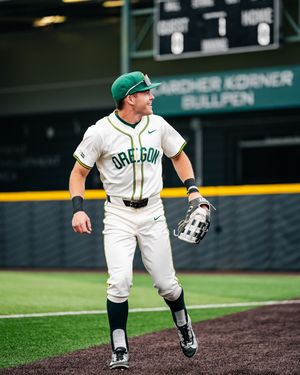Oregon Ducks baseball player in white uniform and green cap jogging on field during a college baseball game.