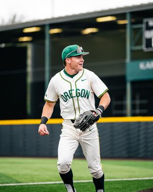 Oregon Ducks baseball player in white uniform and green cap jogging on field with glove during game.