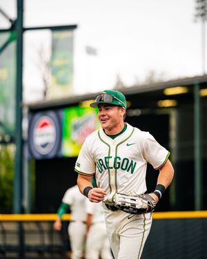 Oregon Ducks baseball player jogging in white uniform and green cap, smiling on the field.