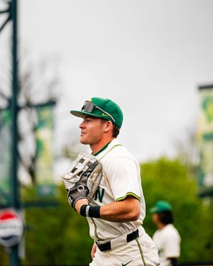 Baseball player in green and white uniform with green cap smiling during an outdoor game.