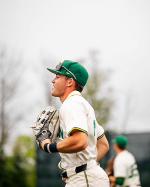 Baseball player in green cap and white uniform holds glove while gazing upward during an outdoor game.