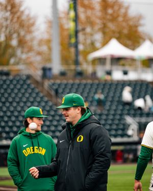 Two Oregon Ducks baseball players in green uniforms chat on the field during an overcast game day.