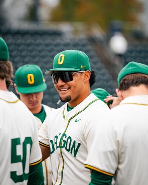 Oregon Ducks baseball players in green and yellow uniforms smiling together in the dugout during a game.
