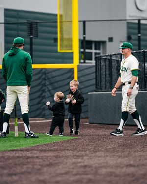 Two toddlers interact on a baseball field while players in green uniforms watch during a team event.