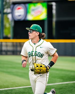 Oregon Ducks baseball player jogging in cream uniform with green cap and Marucci glove on outdoor field.