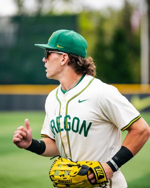 Oregon Ducks baseball player in green cap and white uniform jogging on field holding a yellow Marucci glove.
