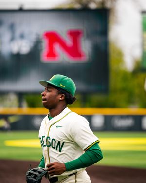 Oregon Ducks baseball player in green uniform and cap on field with Nebraska Cornhuskers scoreboard in background.