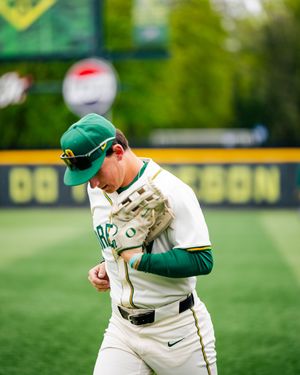 Oregon Ducks baseball pitcher in green cap and white uniform walks on field holding glove during game.
