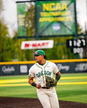 Oregon Ducks baseball player in white and green uniform jogging on field with NCAA Tournament scoreboard in background.