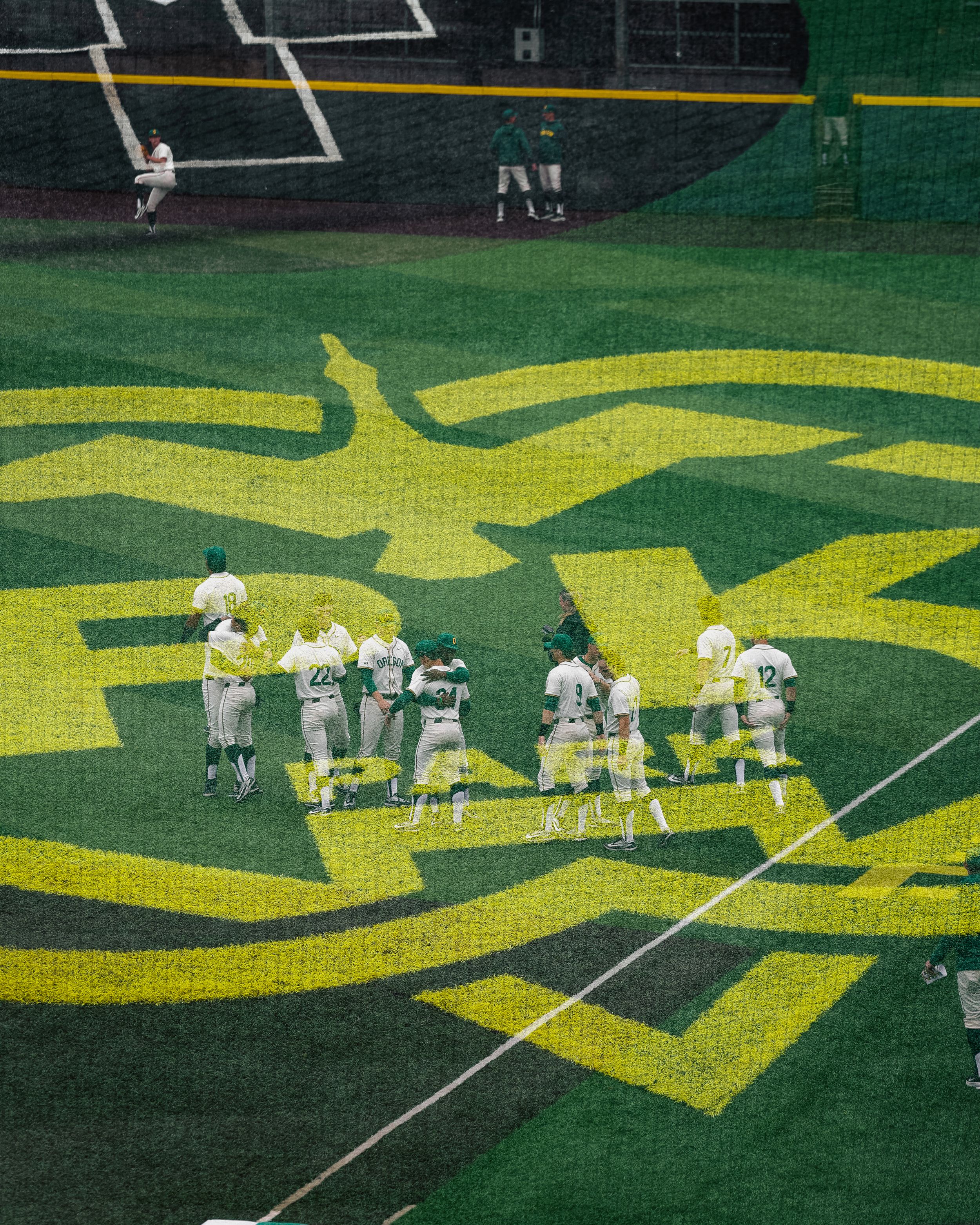 Baseball team huddles on a green field with a large yellow duck logo painted on the grass at a stadium.