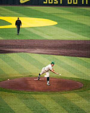 Baseball pitcher mid-throw on mound at Nike-sponsored stadium with striped green outfield and umpire in background.