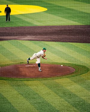 Baseball pitcher in white and green uniform mid-throw on pitching mound during a college baseball game.