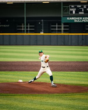 College baseball pitcher in white and green uniform delivering pitch from mound at professional stadium.