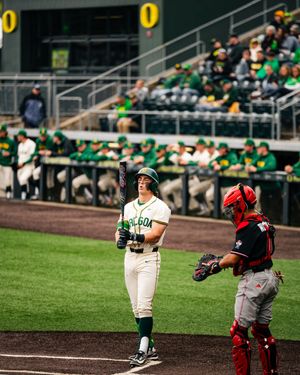 Oregon Ducks baseball player smiles at home plate holding bat next to opposing team red-uniformed catcher during college game.