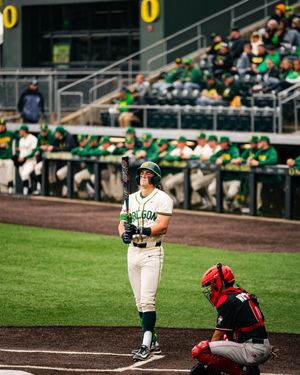 Oregon Ducks batter stands ready at home plate holding bat while red-uniformed catcher crouches during college baseball game.
