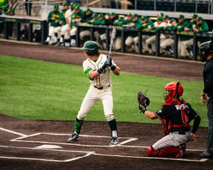 College baseball batter in green uniform at home plate with catcher in red gear during a game.