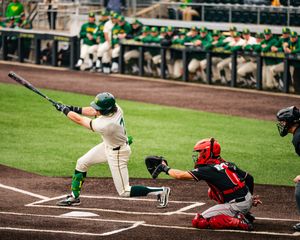Oregon Ducks baseball batter swings at pitch while red-uniformed catcher crouches behind home plate.
