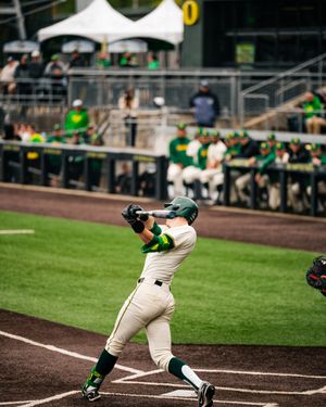 Oregon Ducks baseball player in green and yellow uniform swings bat during a college game at a packed stadium.