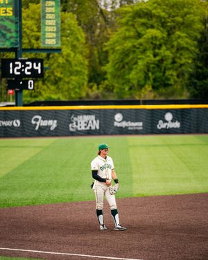 Oregon Ducks baseball player stands on base at PK Park with scoreboard showing 12:24 in background.