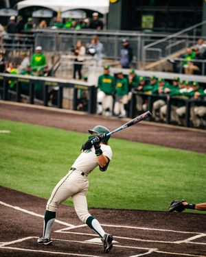 Baseball batter in white and green uniform swings at pitch during college game with dugout and fans in background.