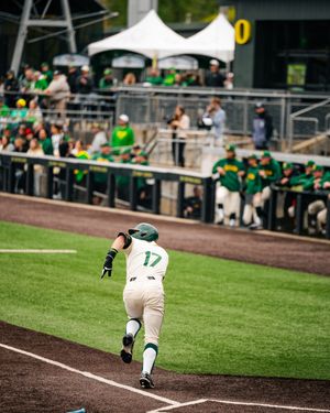 Baseball player #17 in green and white uniform runs the bases during a college baseball game with fans in background.