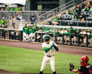 Baseball batter in green uniform stands ready at plate with catcher behind him at a collegiate baseball game.