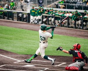 Baseball player #53 in green uniform swings bat at home plate with catcher in red gear behind him.