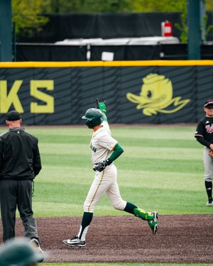Oregon Ducks baseball player rounds the bases after a home run at PK Park, with an umpire and opponent nearby.