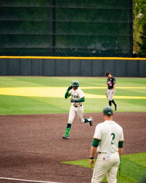 Baseball player in green uniform rounds the bases after a hit during a college baseball game on a well-maintained field.
