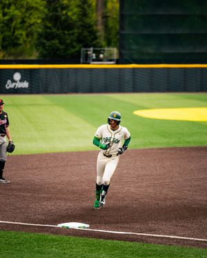 Oregon Ducks baseball player in green uniform rounds third base during a college baseball game at a green stadium.
