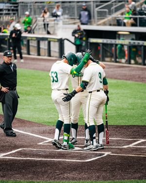 College baseball players in green and white uniforms celebrate at home plate after a successful hit during a game.