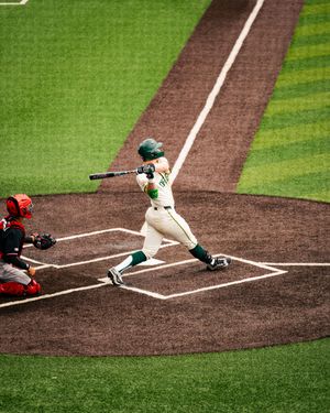 Baseball batter in green uniform swings at pitch while catcher crouches behind home plate on a manicured field.