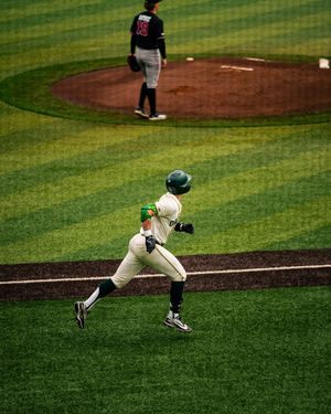 Baseball player in green helmet runs bases as pitcher stands on mound during a college baseball game.