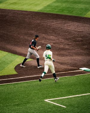 Baseball player in green helmet runs to first base as fielder in black uniform prepares to make the catch.
