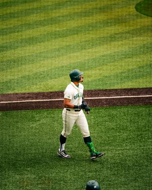 Oregon Ducks baseball player in white uniform and green helmet walks on field during a college baseball game.