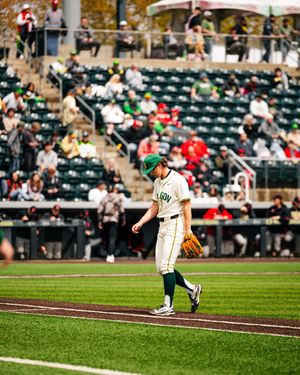 A baseball pitcher in green and white uniform walks on the field during a game at a stadium with fans in the stands.